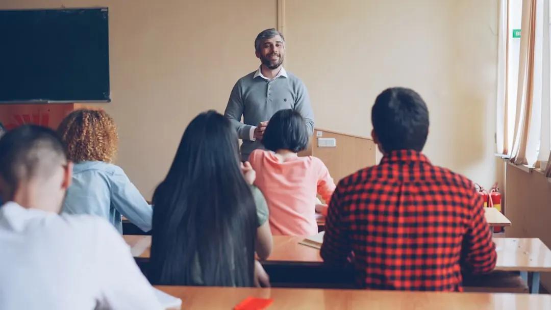 A teacher standing at the front of a classroom while students at desks listen and take notes