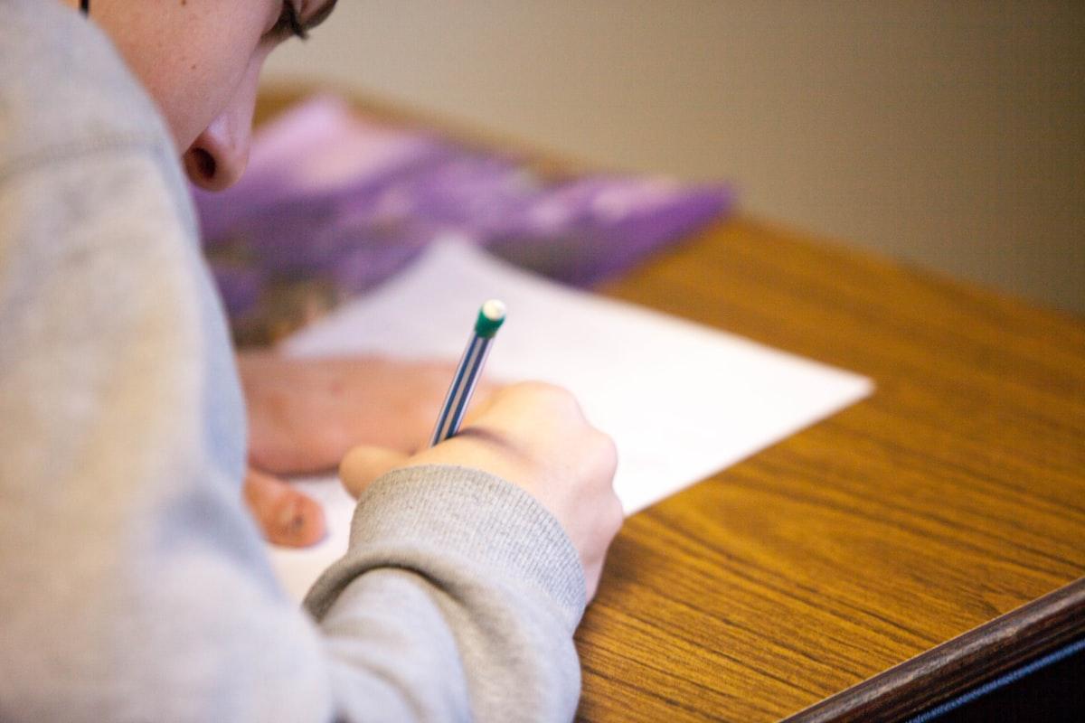 A teacher sits at a desk reviewing a printed student essay with a pen in hand